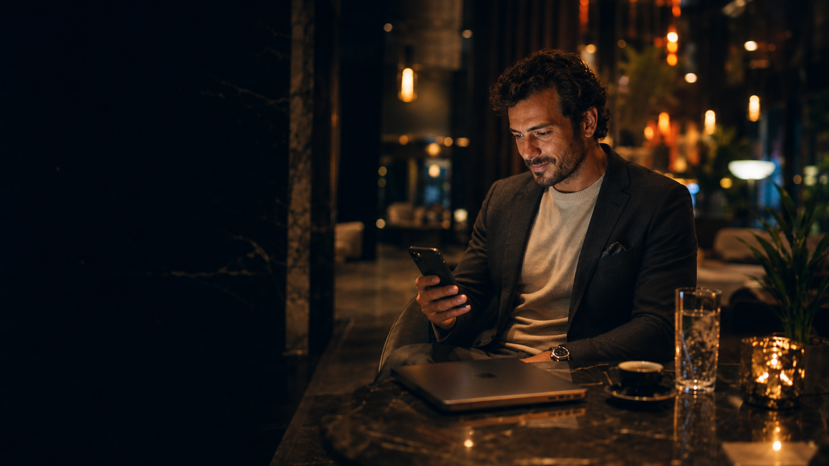 Man at a warm candle-lit café looking at his phone, city lights glowing behind him